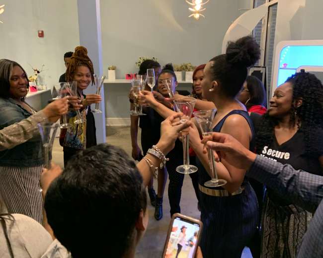 A group of people stands together, holding up glasses in a toast while smiling and celebrating indoors.