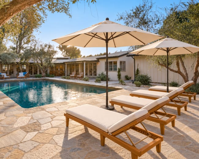 The image shows a tranquil outdoor pool area with lounge chairs and umbrellas surrounded by greenery and a spacious stone patio.