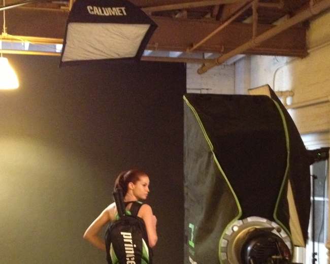 A model poses in a photography studio while wearing a backpack and standing next to a large softbox light.