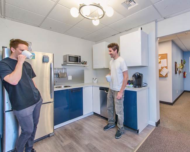 Two men are standing in a kitchen area with a refrigerator, microwave, and coffee maker while one drinks from a cup and the other holds a mug.
