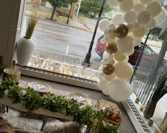 The interior showcases a decorative setup for a celebration, featuring a table with greenery, white and gold balloons, and illuminated letters spelling "MARRY ME" against a window view.