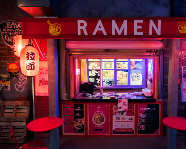A ramen stall with a bright red sign, illuminated lanterns, and a menu displayed in the window.