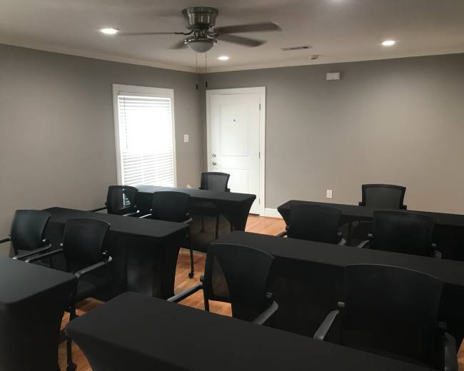 The image shows a conference room with several black tables and chairs arranged in a layout suitable for meetings, with a door and windows visible.