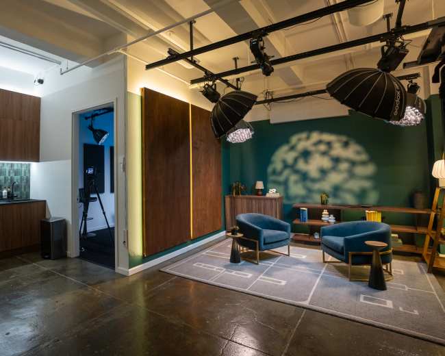 The image shows a modern studio space featuring a kitchenette area, a vibrant green wall with soft lighting, and two blue chairs arranged near a decorative wooden console table.