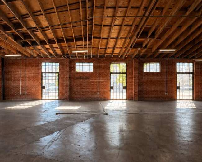 The image shows an empty industrial space with exposed brick walls, a high wooden ceiling, and large windows allowing natural light to enter.