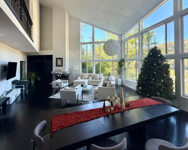 A spacious living area features large windows, a dining table with a red table runner, modern furniture, and a Christmas tree in the corner.