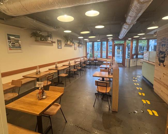 The image shows a spacious cafe interior with wooden tables and chairs arranged along the walls, and a row of lights hanging from the ceiling.