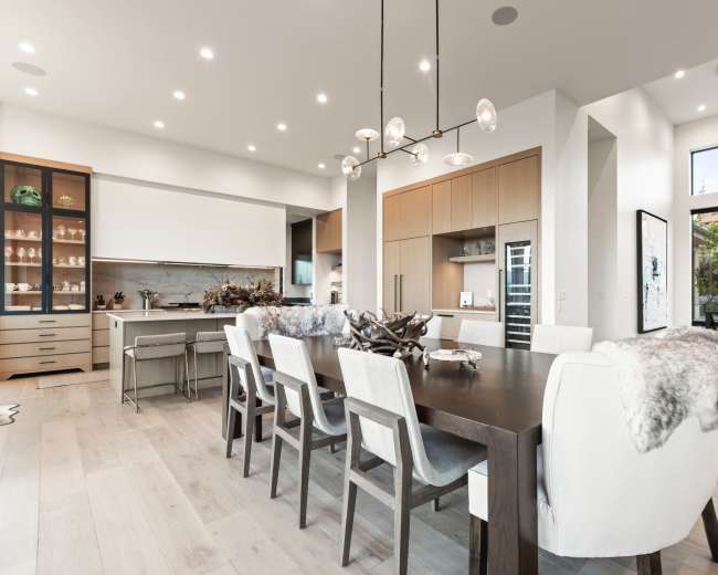 A modern dining area features a large wooden table surrounded by upholstered chairs, with a pendant light overhead and large windows letting in natural light.