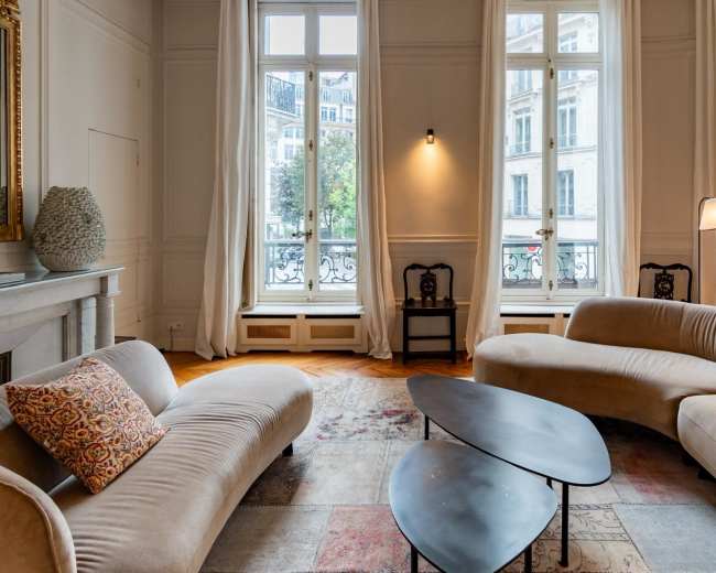 A modern living room features a curved beige sofa, a dark coffee table, and large windows allowing natural light to fill the space.
