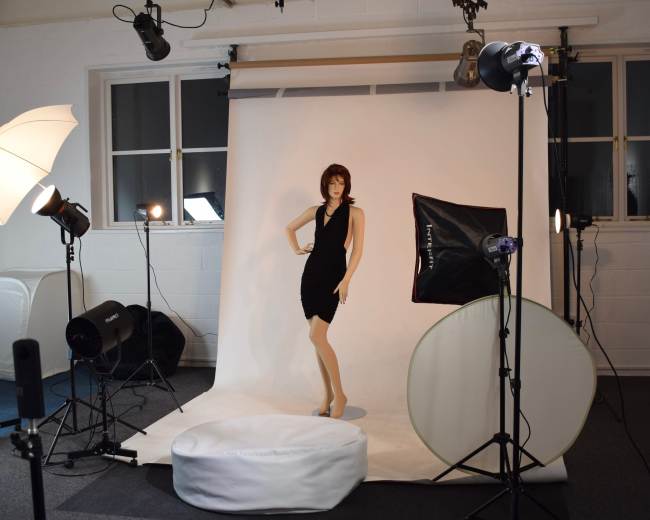 The scene features a mannequin dressed in a black dress, positioned on a circular platform in a photography studio surrounded by various lighting equipment.