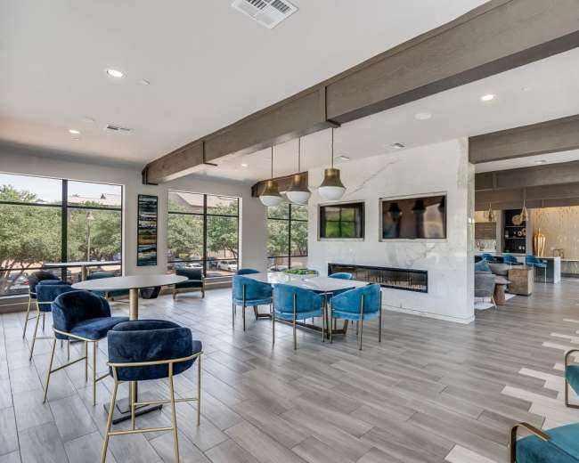 A modern common area with blue chairs, a fireplace, and large windows revealing trees outside.