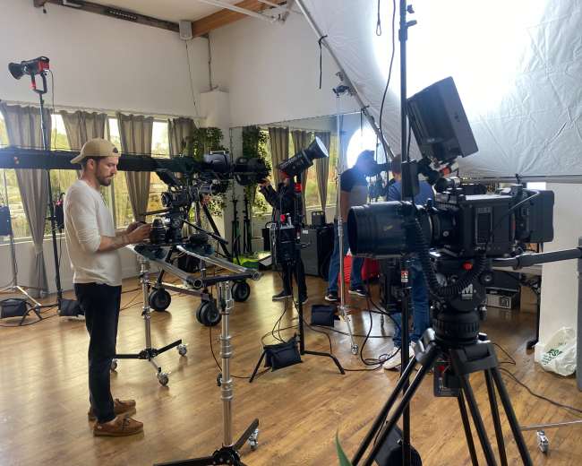 A film crew sets up equipment in a well-lit studio with multiple cameras and lighting setups.