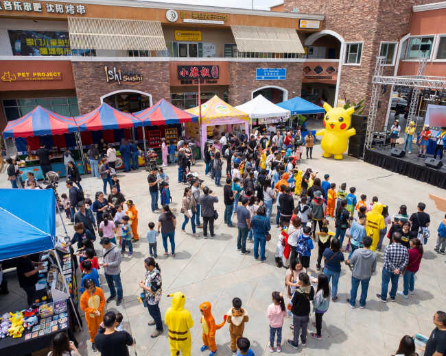 A large crowd gathers in an outdoor plaza filled with colorful tents and a stage, featuring characters like Pikachu while vendors sell various items.