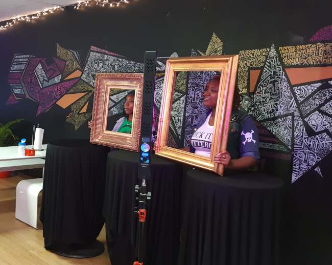 A photographer sets up a camera and lights to capture subjects posing with ornate frames in a vibrant studio adorned with artwork.