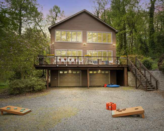 A two-story house with large windows and a deck overlooks a gravel parking area, featuring a pair of cornhole boards and a blue inflatable float nearby.