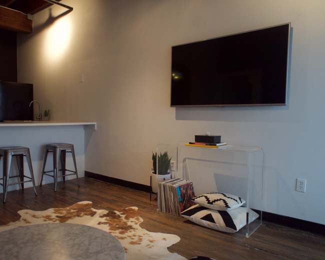 The image shows a modern living space featuring a television mounted on the wall, a clear coffee table with books, a bar counter with two stools, and a cowhide rug on a wooden floor.