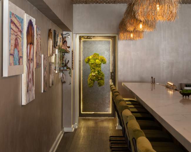 The image shows a narrow restaurant corridor featuring artwork on the walls, a long bar with green stools, and a distinctive door adorned with moss.