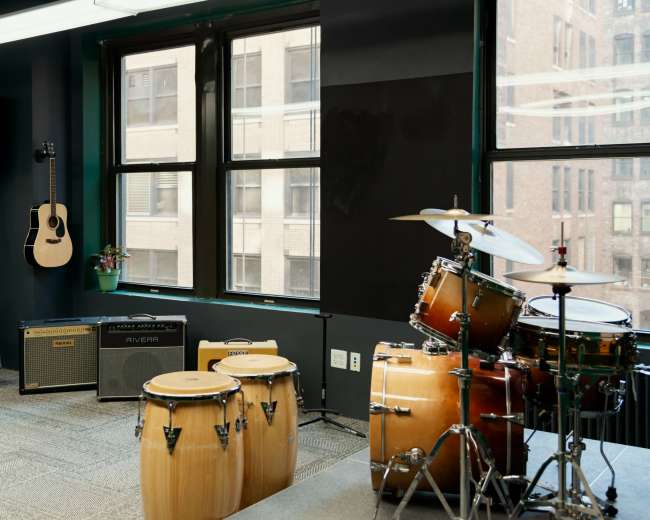 The image shows a music studio with a drum set, conga drums, an acoustic guitar on the wall, and amplifiers beside large windows.