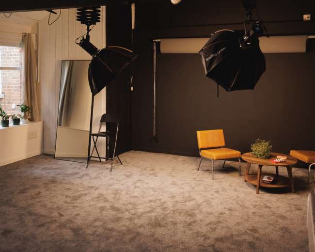 The image shows a spacious photo studio with a dark backdrop, two softbox lights, a mirror, two chairs, and a small table on a carpeted floor.