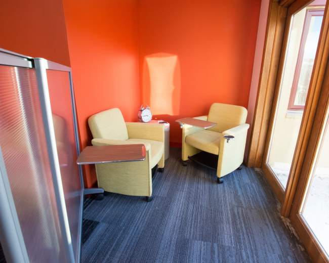 A small seating area with two yellow chairs and a wooden table against a bright orange wall, with a clock on the table and large windows letting in natural light.