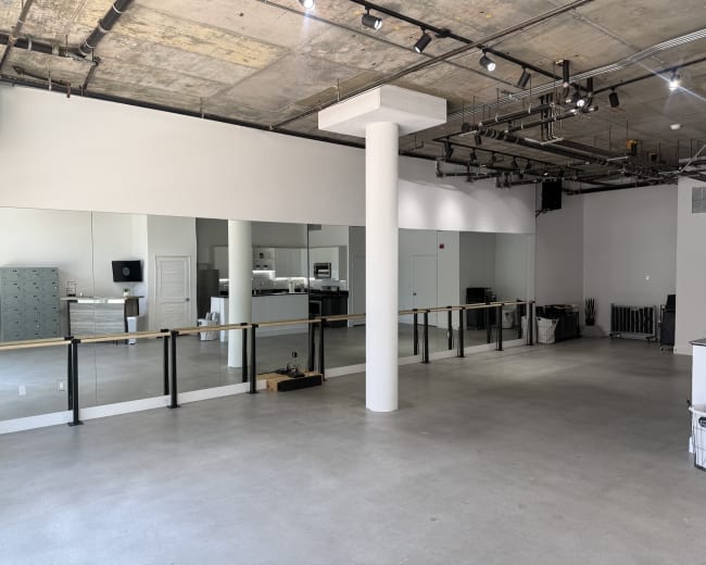 The image shows a spacious, minimalist studio with large mirrors lining one wall and various furnishings, including a counter and storage units, visible in the background.