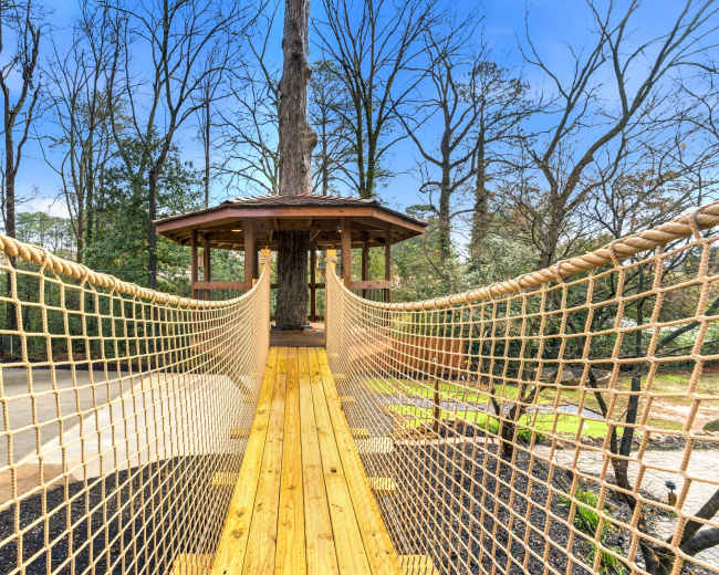 The image shows a wooden suspension bridge with a rope railing leading to a gazebo surrounded by trees.
