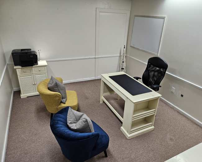A small office space with a white desk, a black office chair, two armchairs, a printer, and a whiteboard on the wall.