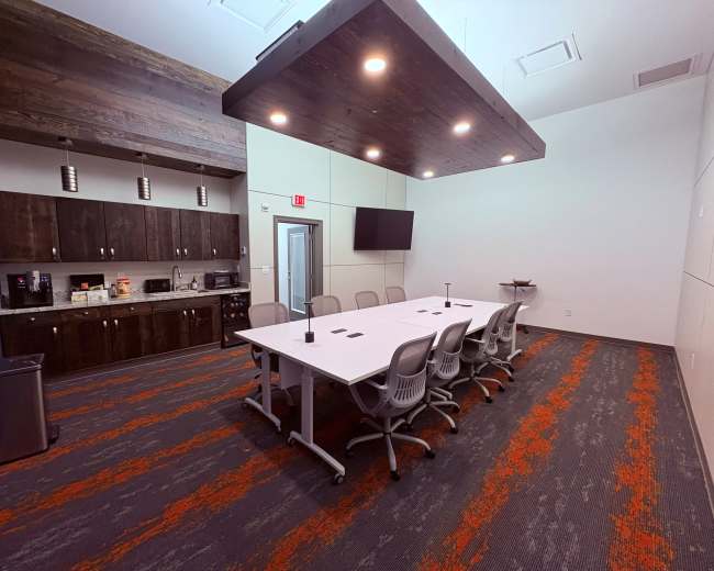 The image shows a modern conference room with a long white table surrounded by chairs, a kitchen area, and a wall-mounted screen.
