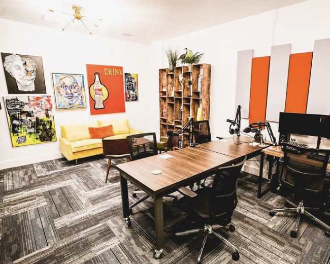 The image shows a modern workspace featuring a sofa, artwork on the walls, a wooden bookshelf, and desks equipped with microphones and computers.