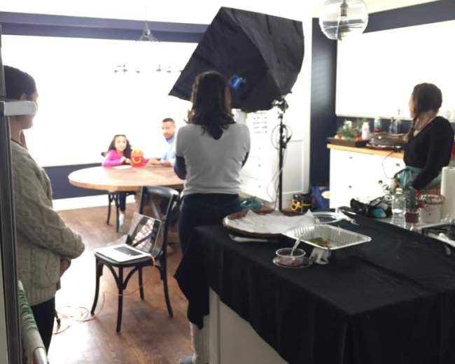 A film crew is set up in a kitchen, with one person adjusting a light while others are positioned around a wooden table.