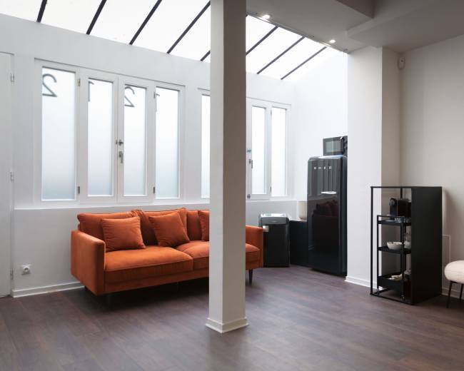 The image shows a modern, minimalist room featuring an orange sofa, a black refrigerator, and a shelving unit against white walls with large windows.