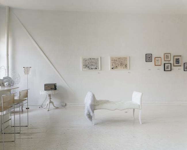 A bright, minimalist art gallery features white walls, a bench with a fur throw, and several framed artworks hanging on the walls.
