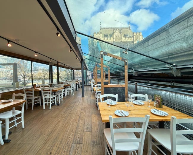 The image shows an indoor dining area with wooden tables and white chairs, featuring large windows that offer a view of a building outside.