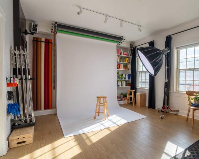 The image shows a well-lit photography studio with a white backdrop, a wooden stool, and a variety of colorful backdrops and props stored along the wall.