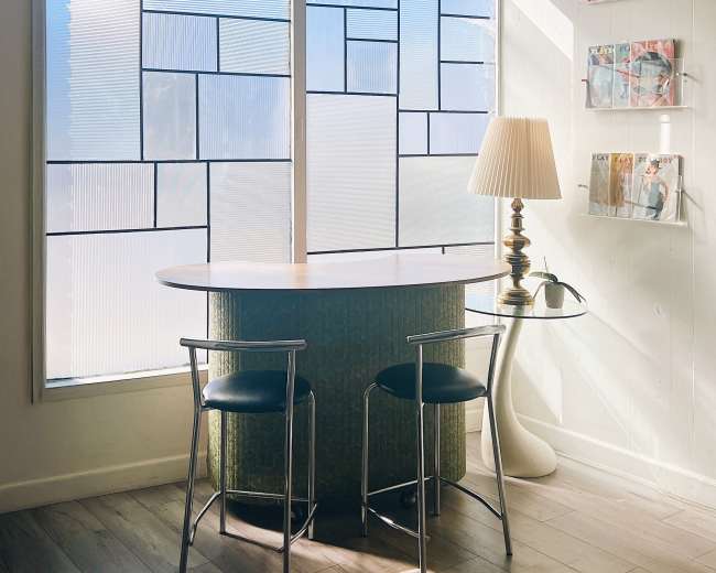 A small, modern workspace features a round table with two chairs and a lamp, illuminated by natural light streaming through large, patterned windows.