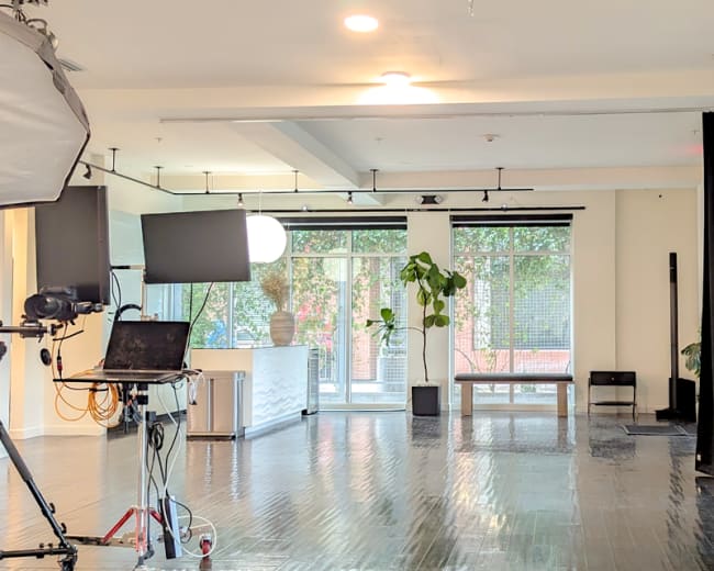 A bright, open studio space with large windows, dark curtains, and a video setup including a camera and monitor.