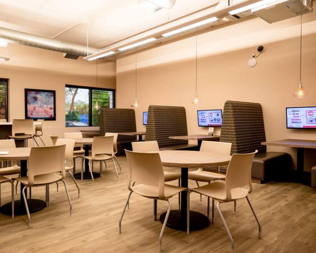 The image shows a spacious, modern café interior with several tables and booth seating, illuminated by overhead lighting and featuring electronic displays on the walls.