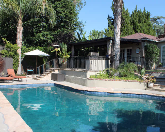 The image shows a backyard with a swimming pool, surrounded by tropical plants and a patio area with lounge chairs and an umbrella.