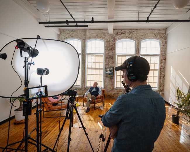 A person wearing a headset stands in front of a camera setup, while another person sits on a couch in a well-lit, modern room with large windows and wooden floors.