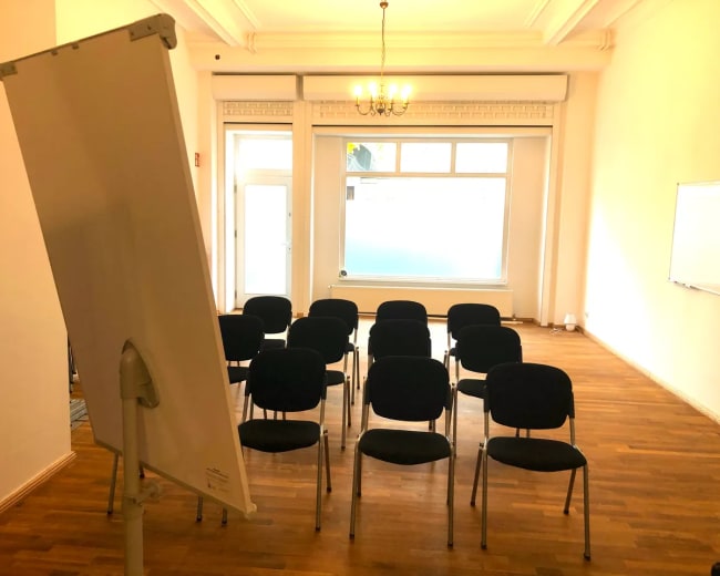 A spacious room with wooden flooring contains several chairs arranged in rows facing a flip chart and a large window.