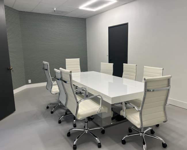 A modern conference room features a large white table surrounded by six white rolling chairs and a dark door on one wall.