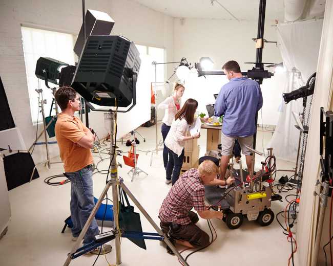 A group of five people work together in a studio setting, setting up equipment and adjusting a camera for a photoshoot.