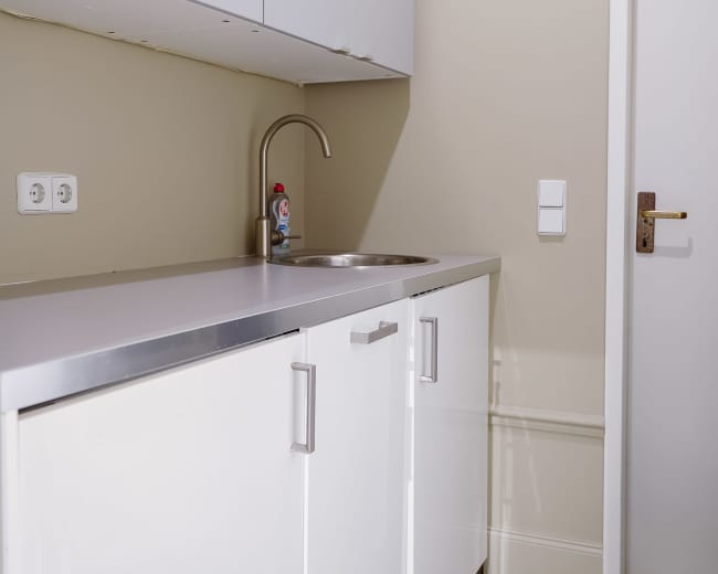 The image shows a small kitchen area with a stainless steel sink and a countertop beneath white cabinets, adjacent to a closed door.