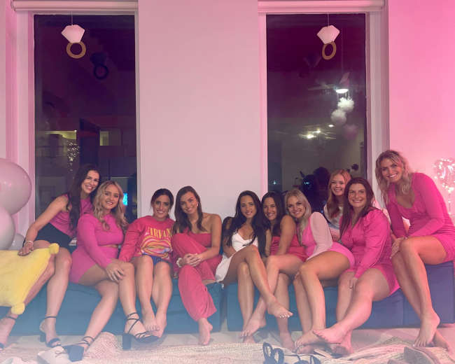 A group of ten women in matching outfits sits on a couch in a well-lit room with large windows and decorative elements.