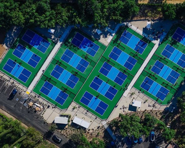 Aerial view of multiple tennis courts surrounded by trees and parked cars.