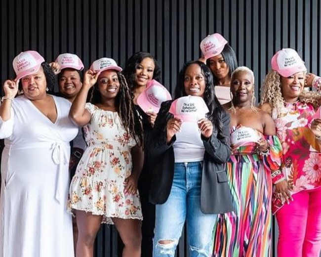 A group of eight women, wearing coordinated hats, pose together in front of a dark wooden wall.