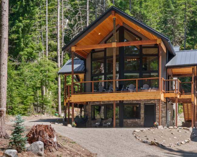 A modern cabin with large windows and a wooden deck is surrounded by tall pine trees and a gravel pathway.