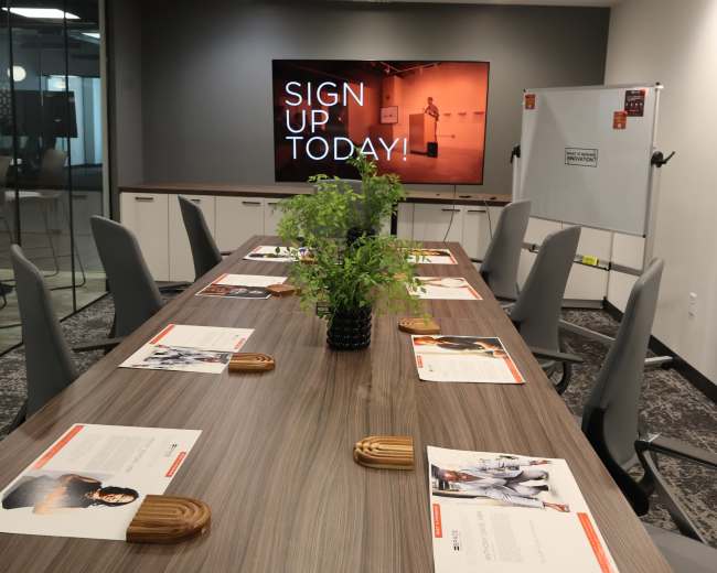 A modern conference room features a large table with chairs, promotional materials laid out, and a screen displaying the message "SIGN UP TODAY!" against a gray wall.
