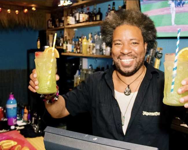 A bartender holding two colorful cocktails with fruit garnishes stands behind a bar filled with various bottles.
