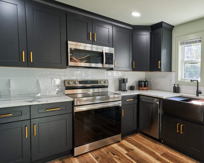 The image shows a modern kitchen with dark cabinetry, stainless steel appliances, and a marble countertop featuring a sink and window.